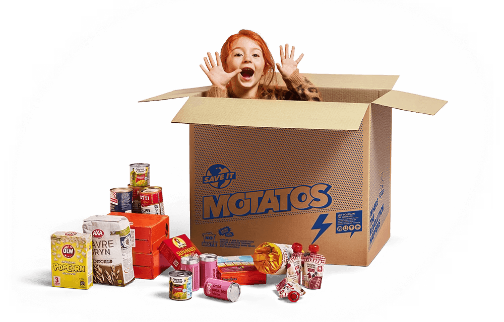 A smiling child with red hair pops out of a large cardboard box labeled "Motatos" with hands raised in excitement. The box has the slogan "Save It" and is surrounded by various food items including popcorn, oat cereal, canned goods, snacks, and drinks.